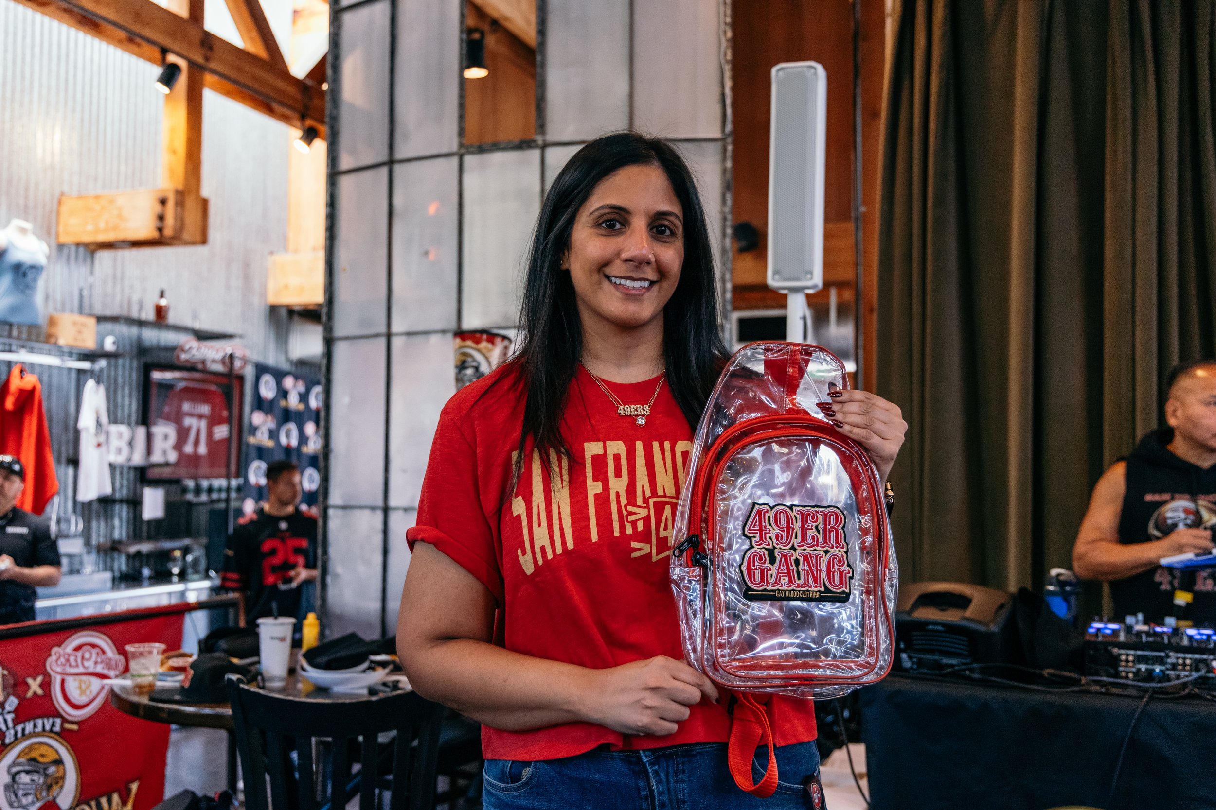 A woman wearing a red San Francisco 49ers shirt on stage at an indoor event, holding a transparent bag with a 49ers Gang logo, with people and merchandise in the background.