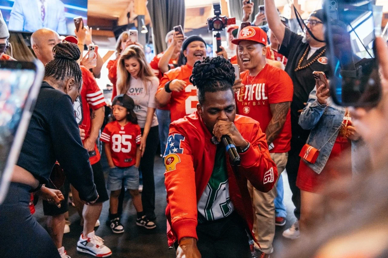 A group of San Francisco 49ers fans celebrating and enjoying a karaoke session inside a venue. The man in front is singing into a microphone with his eyes closed, wearing a red 49ers jacket and shirt, surrounded by smiling and cheering fans in team g