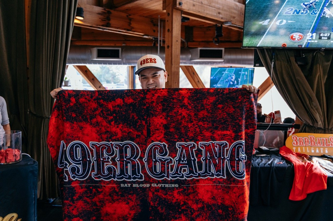 A man wearing a white cap with '49ERS' on it is holding a red and black blanket that says '49ER GANG' in a sports bar or restaurant. There are TVs showing football games, and some tables with red clothing and signage in the background.