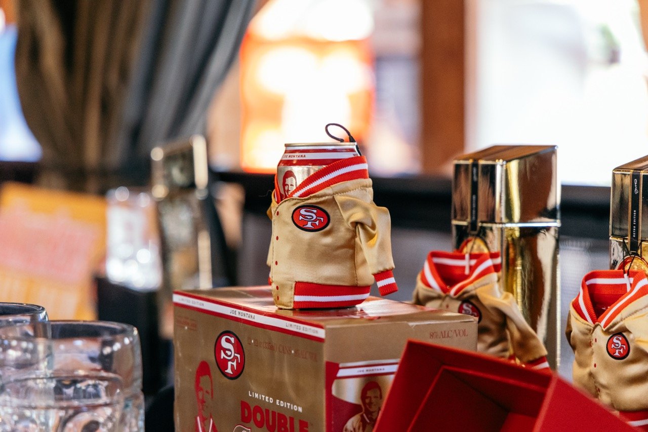 A can of beer dressed in a beige San Francisco 49ers-themed jacket sitting on a cardboard box, with other similar jackets and shiny metallic containers nearby.
