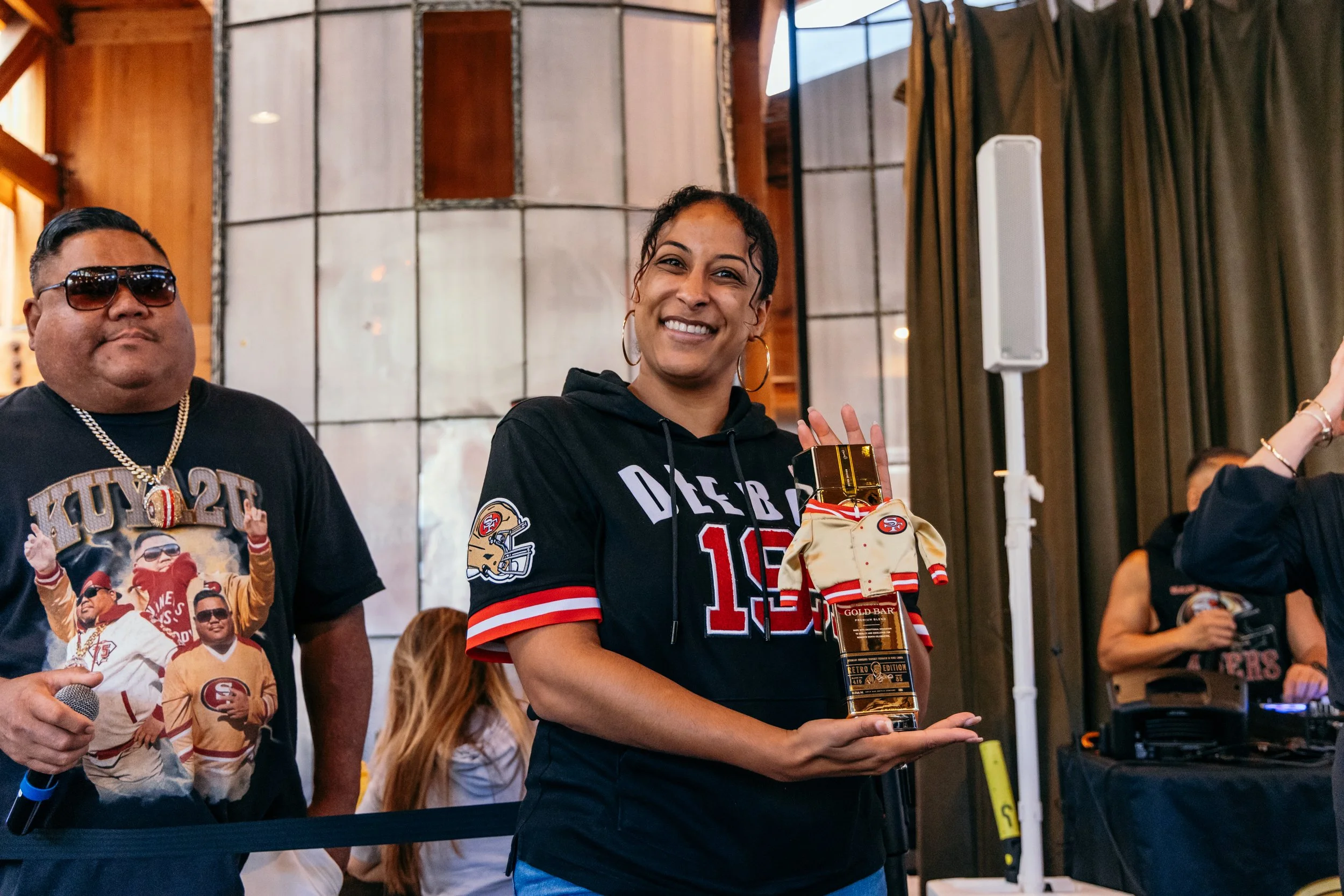 A woman wearing a black hoodie with NFL San Francisco 49ers logo, smiling and holding a boxed gold bar. She is standing in a room with wooden walls and curtains, with other people visible in the background, some wearing San Francisco 49ers apparel.