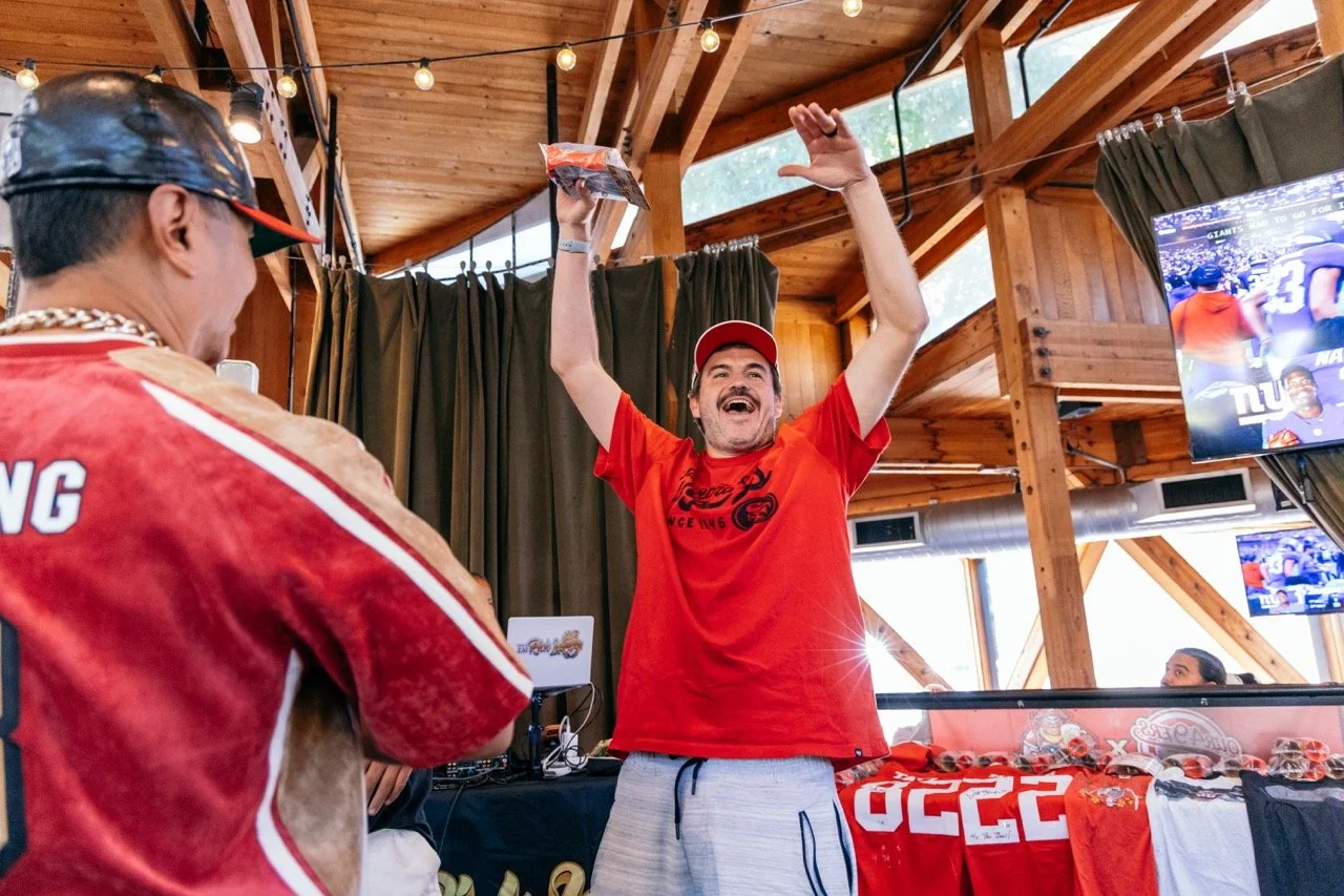 A man in a red t-shirt and red cap celebrating with his arms raised in a wooden indoor space, with another person in a sports jersey in the foreground and multiple television screens showing football games.