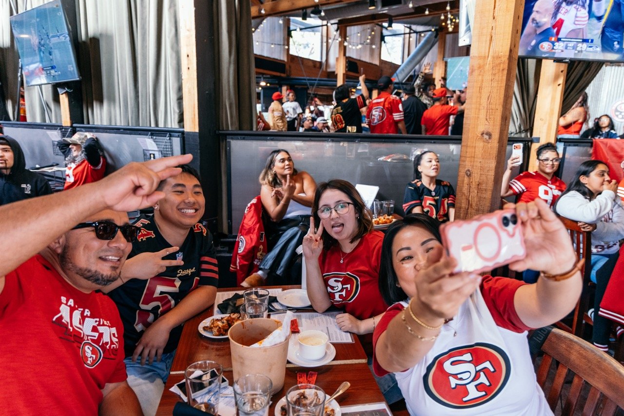 People wearing San Francisco 49ers jerseys and shirts at a sports bar, taking selfies and celebrating a football game.