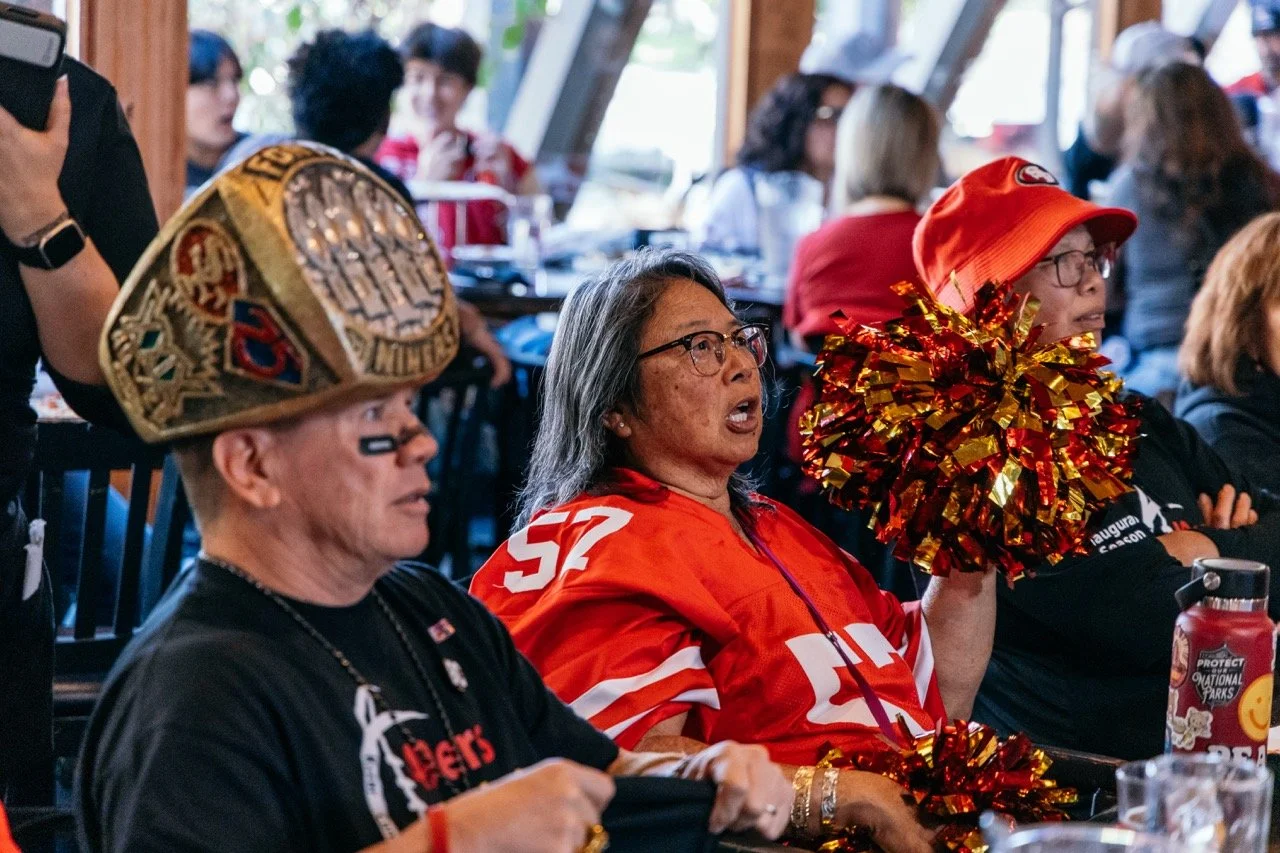 Women at a sports viewing party, wearing San Francisco 49ers team gear and festive hats, some holding gold and red pom-poms.