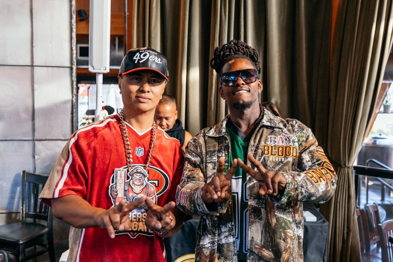 Two men posing together indoors. The man on the left wears a San Francisco 49ers cap, a red 49ers jersey, and a thick chain necklace. The man on the right wears sunglasses, a camouflage jacket with patches, and a green shirt underneath. Both are maki