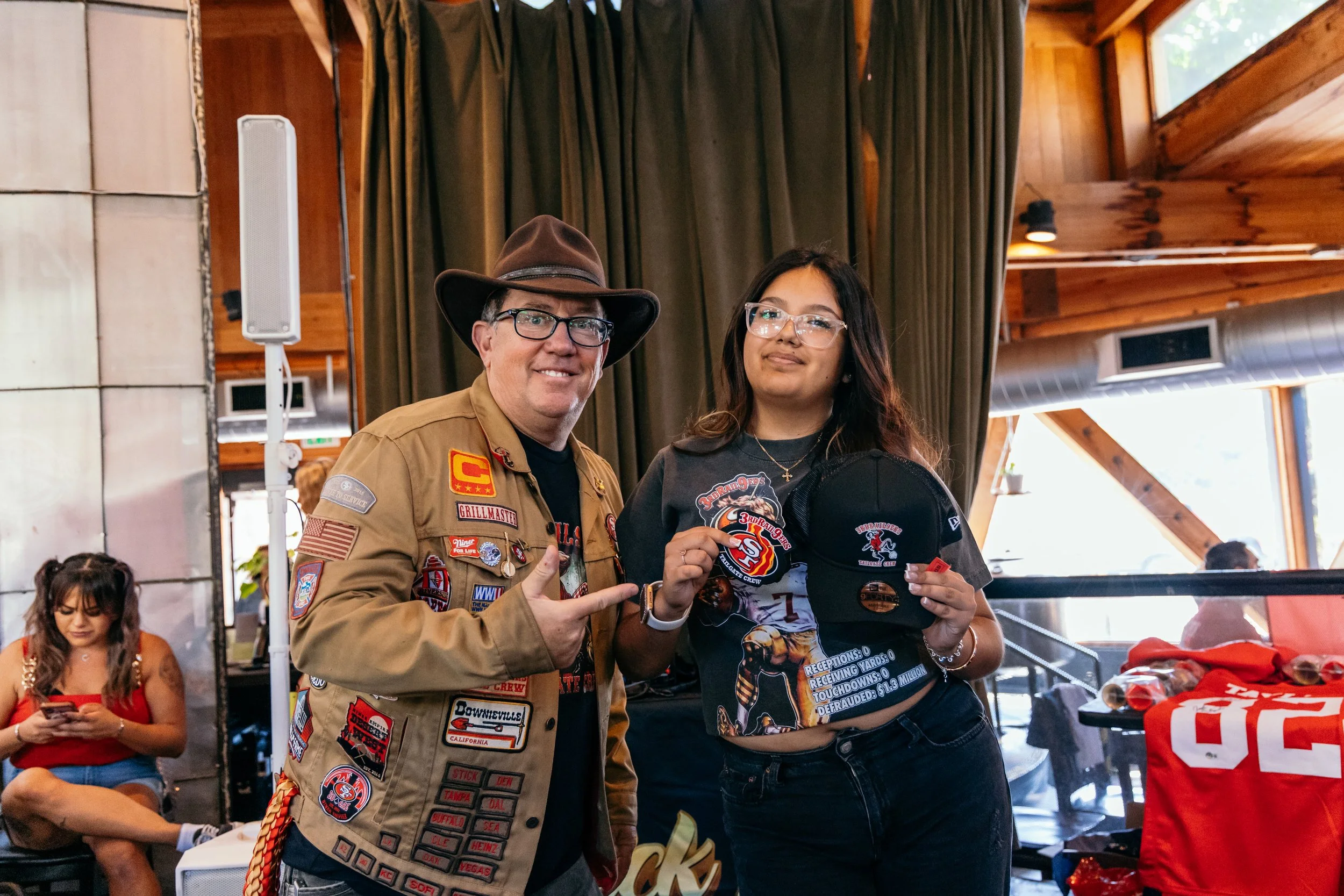 A man wearing a tan jacket decorated with patches and a wide-brimmed hat is standing next to a young woman with long dark hair and glasses. They are indoors with wooden walls and large windows. The woman is holding a small round object, possibly a ba