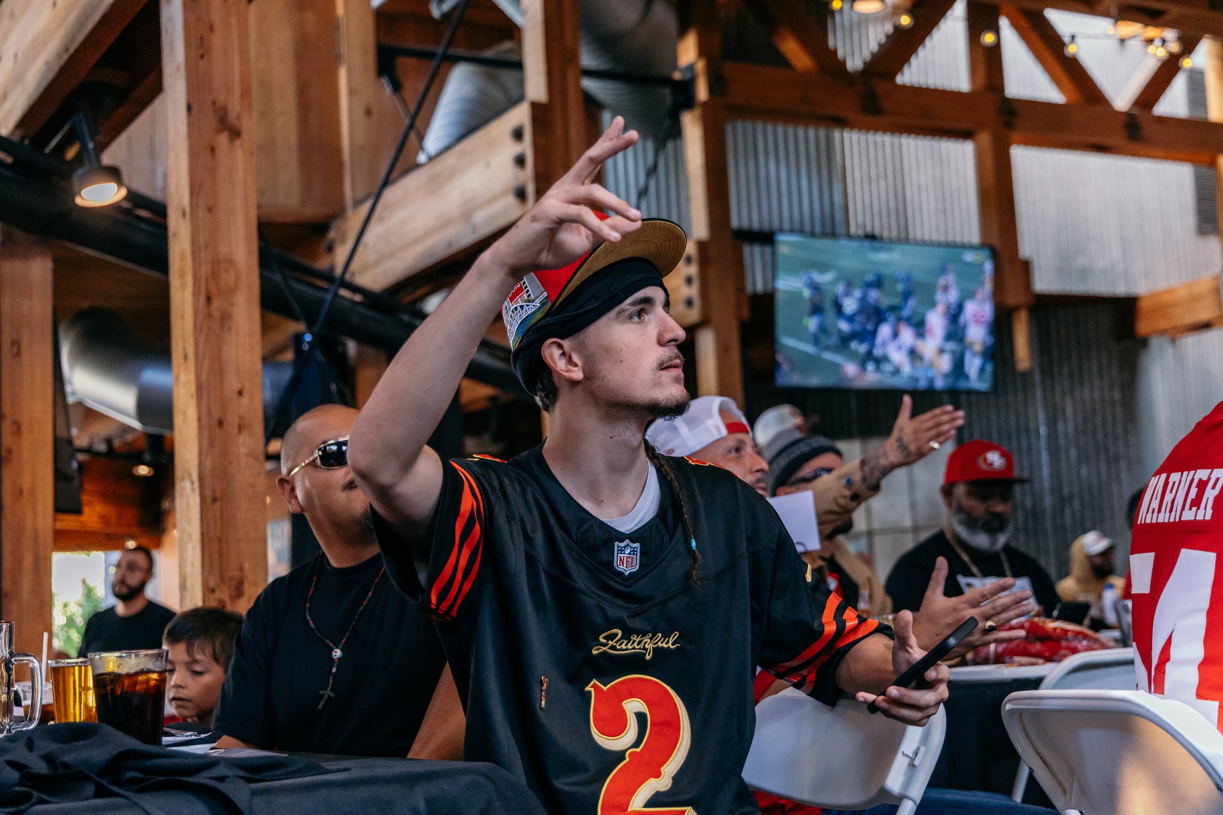 A man wearing a black football jersey with the number 2 and the word 'Faithful' on it, raising his hand, at a sports bar or restaurant during a football game on the TV screen in the background. Others are around him, some wearing football gear.
