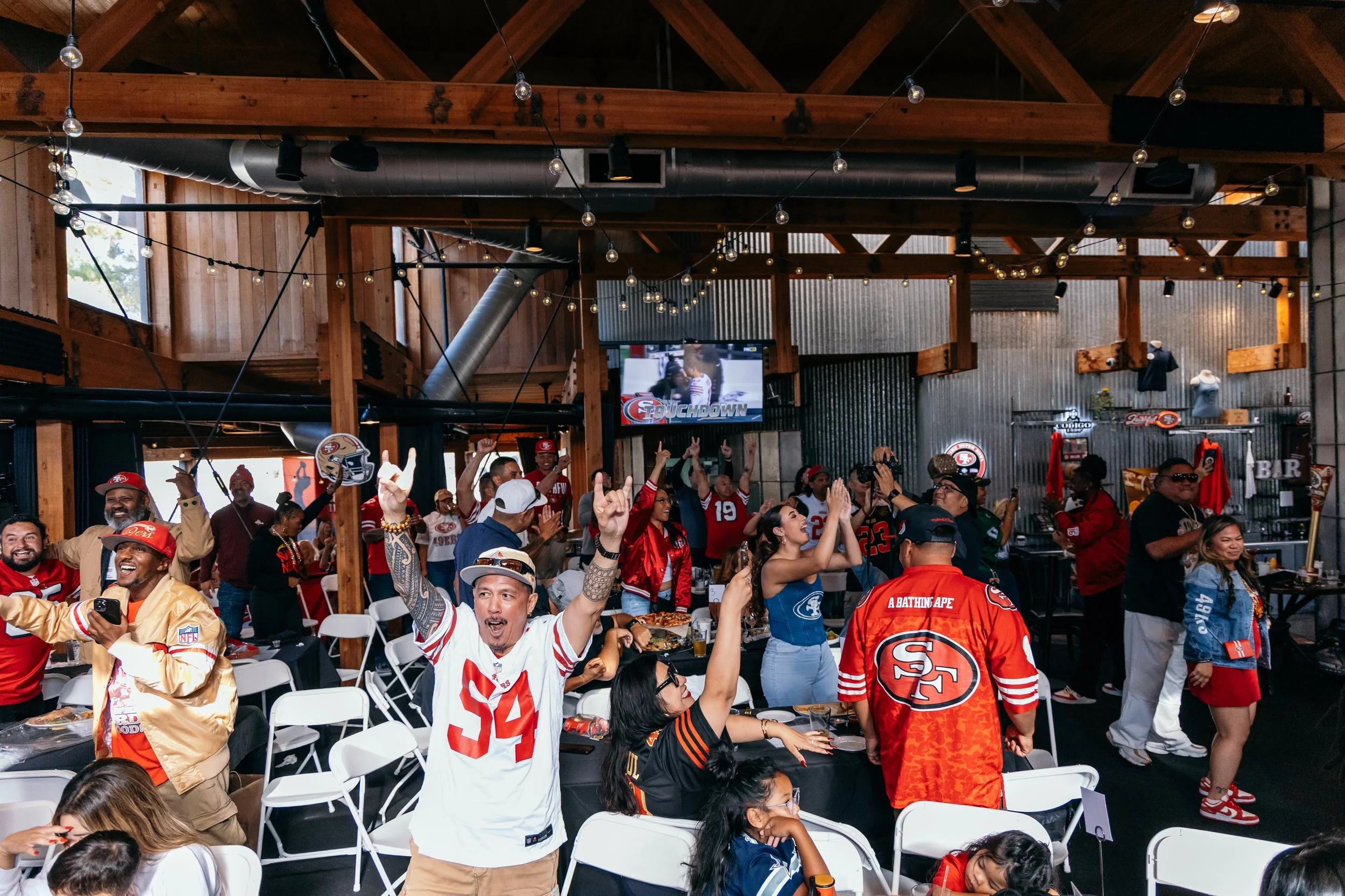 A group of fans celebrating at a party, many wearing San Francisco 49ers and other football team jerseys, inside a rustic-style venue with wooden beams and lighting string. Some are cheering, taking photos, or clapping.