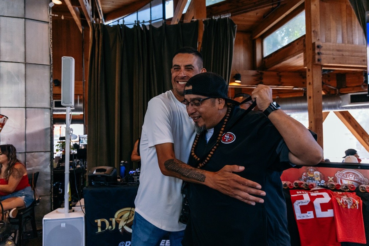 Two men are hugging and smiling inside a wooden-paneled venue with curtains and large windows. One man is wearing a white T-shirt, and the other is wearing a black San Francisco 49ers jersey and a black cap backward. A woman in a red top and shorts i