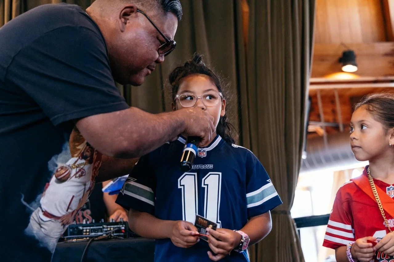 An adult man is speaking into a microphone held close to a young girl wearing glasses and a Dallas Cowboys football jersey. The girl is holding a card and looking at the man. Another girl in a red jersey stands nearby, watching.