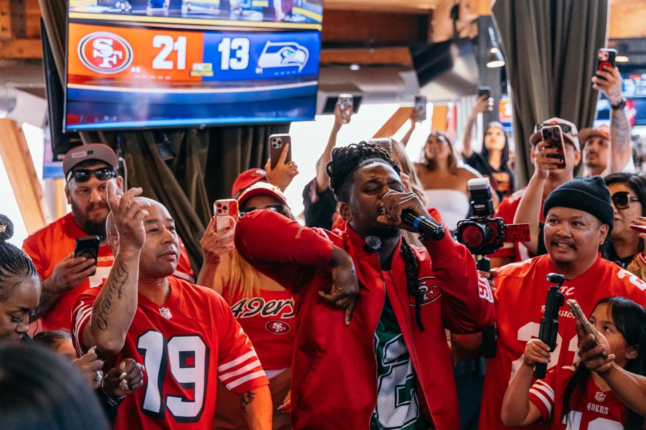 A group of people wearing San Francisco 49ers jerseys watching a game on TV in a bar or restaurant while some are recording the event on their phones. One woman with braided hair is singing into a microphone.