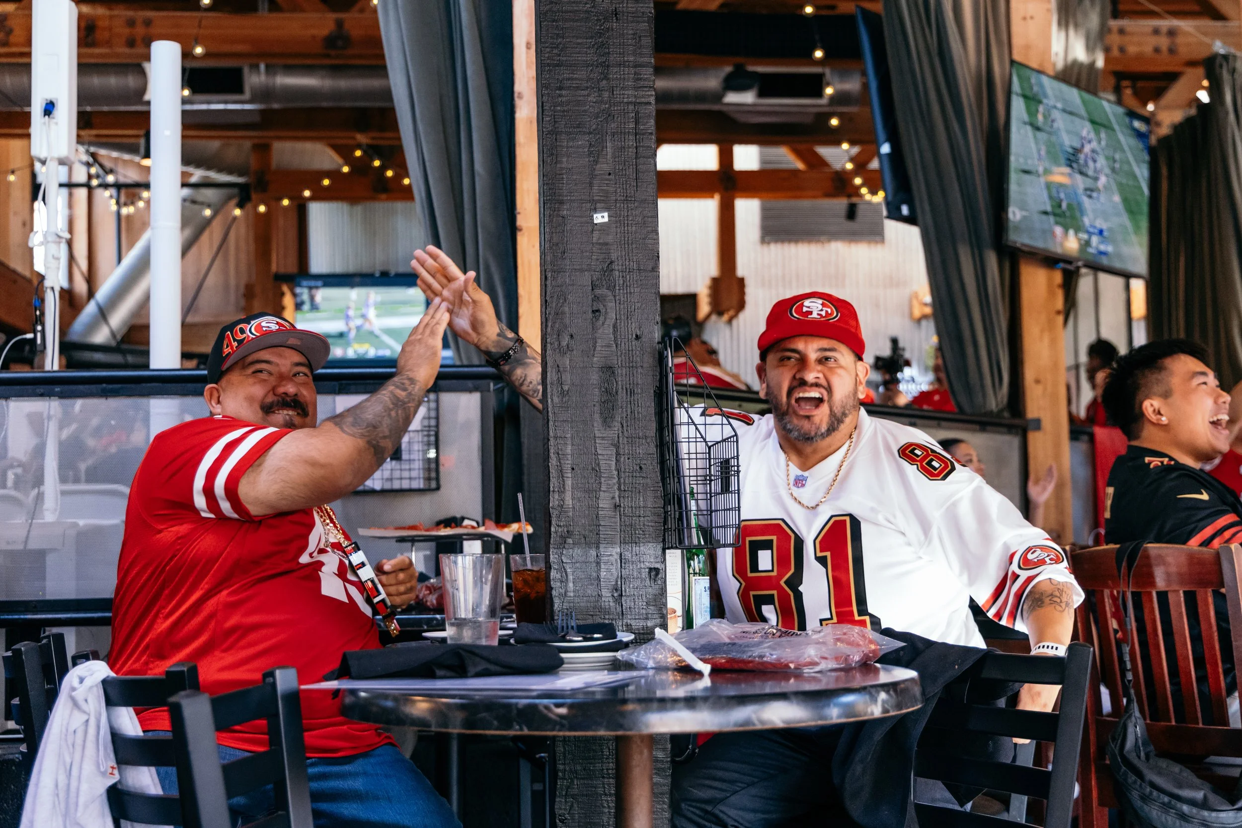 Two men, wearing San Francisco 49ers jerseys and hats, are sitting at a table in a restaurant, high-fiving each other and smiling, with televisions showing football games in the background.