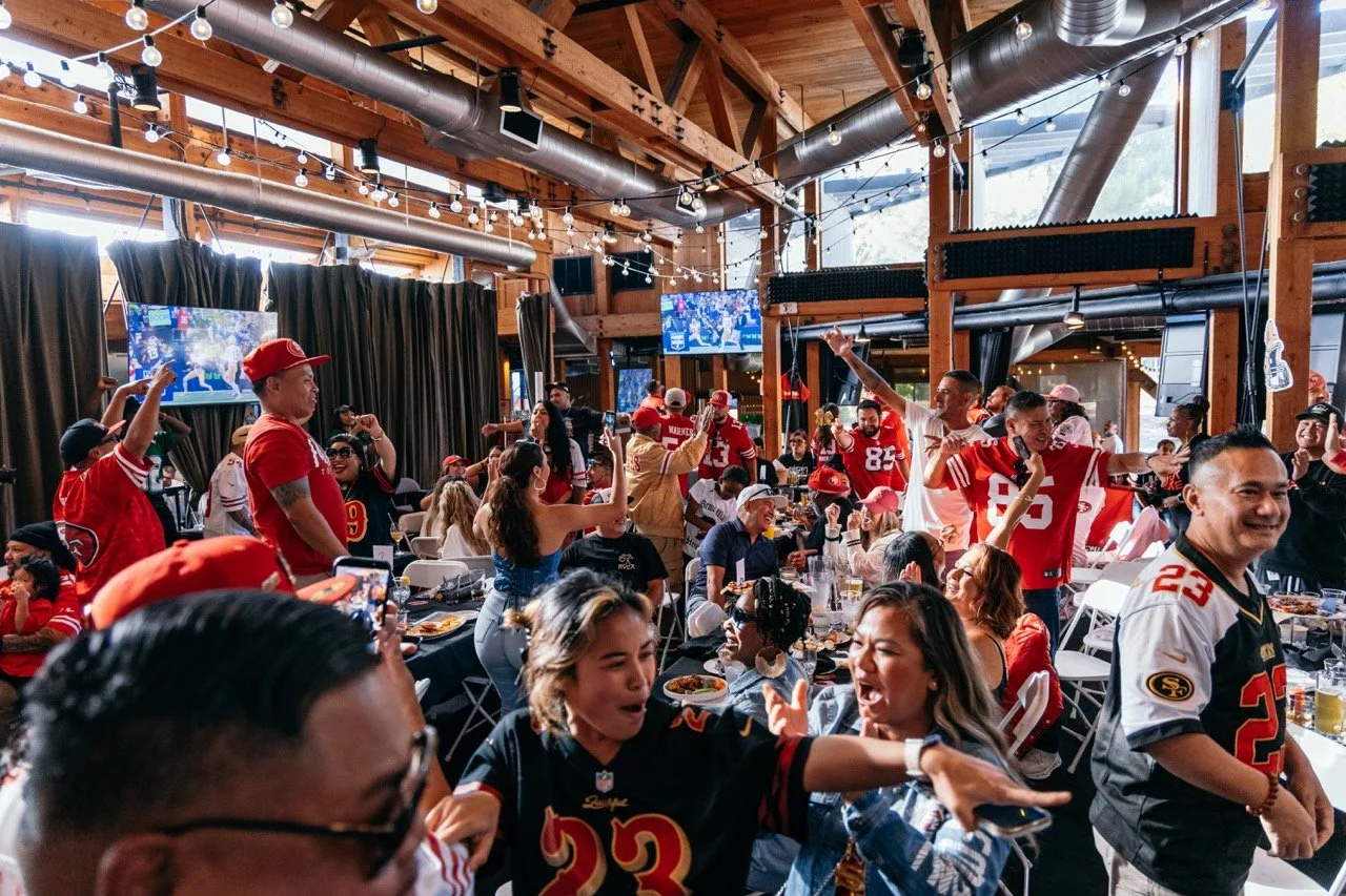 People inside a sports bar or restaurant celebrating, many wearing San Francisco 49ers jerseys and hats, with some raising their hands and cheering, with large TV screens displaying sports games, decorated with string lights and wooden beams.