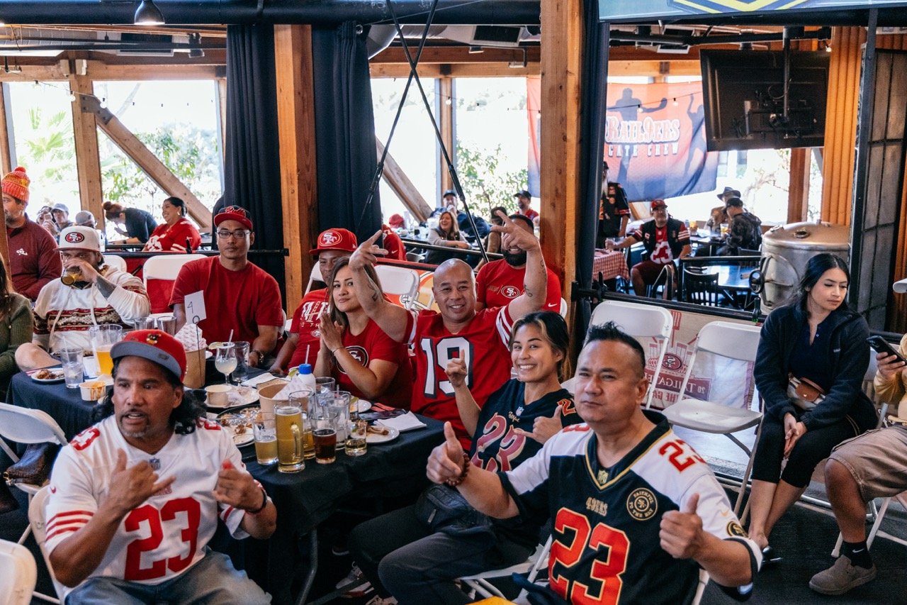 Group of San Francisco 49ers fans at a restaurant, wearing team jerseys and hats, celebrating and socializing while watching a game.