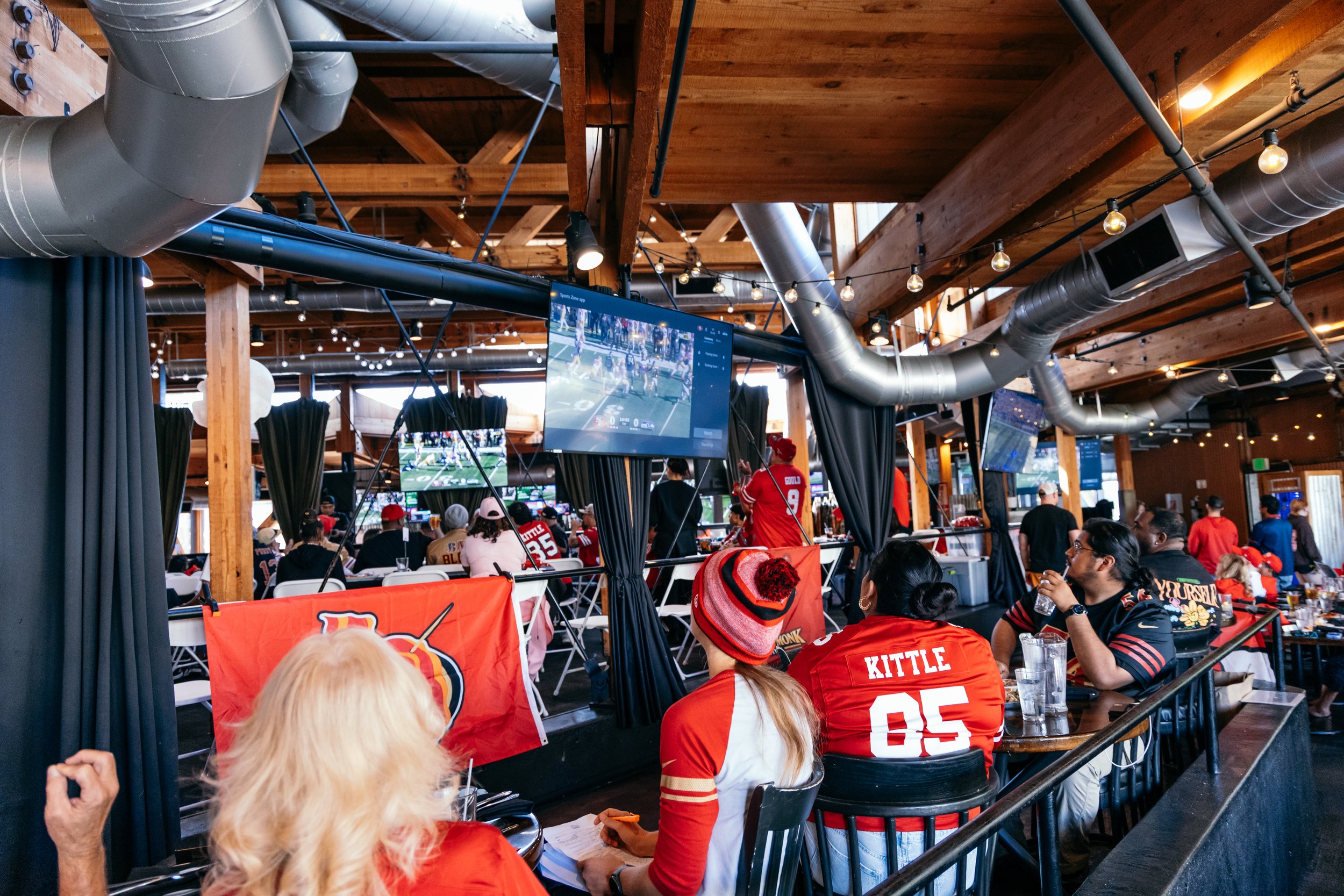 People watching a sports game on multiple large screens at a bar or restaurant, with fans wearing red jerseys, and the interior features exposed wooden beams and industrial ductwork.