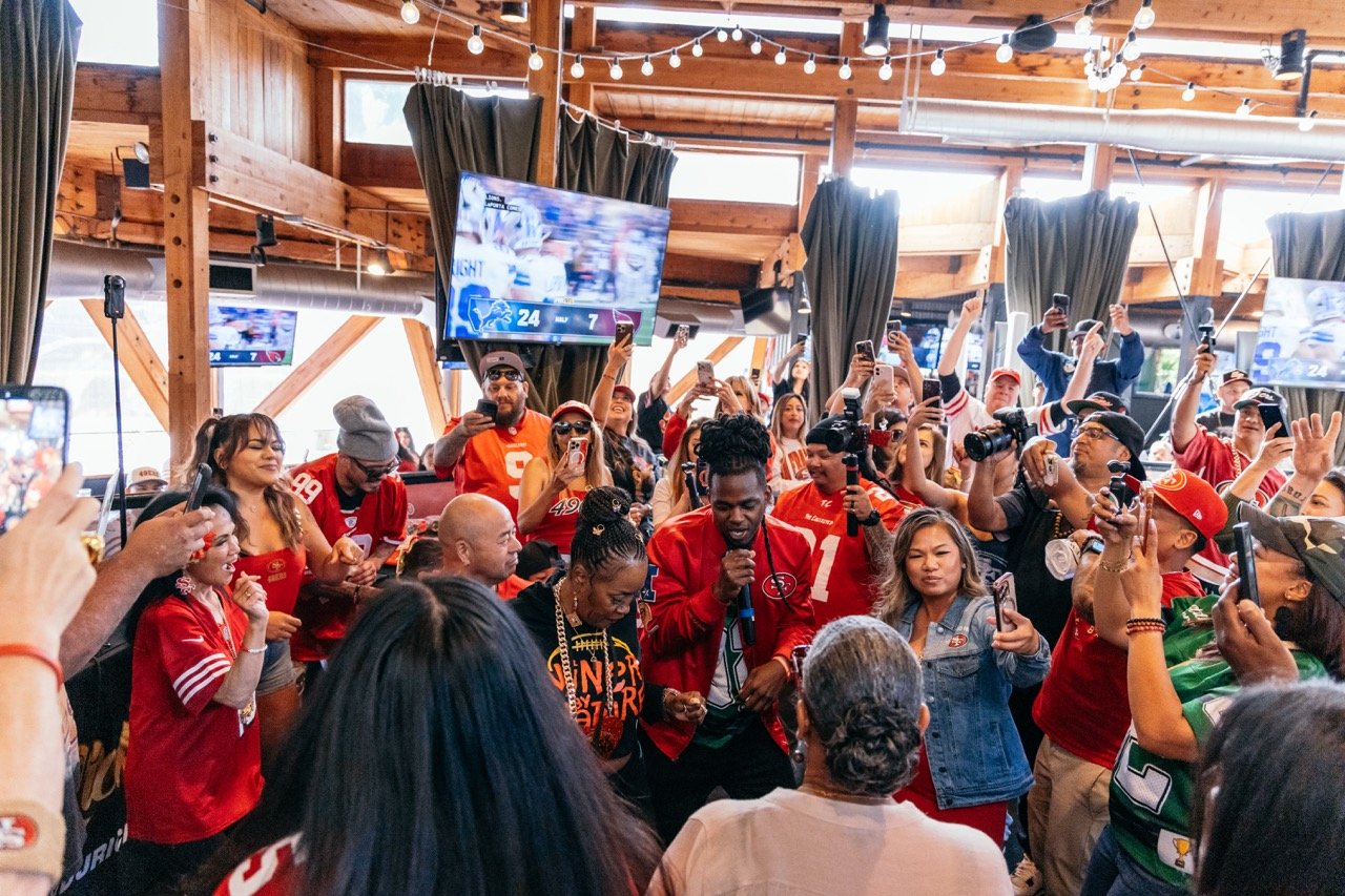 Crowd of NFL fans wearing San Francisco 49ers jerseys and gear inside a wooden sports bar or lounge, celebrating while watching a game on large screens and taking photos.