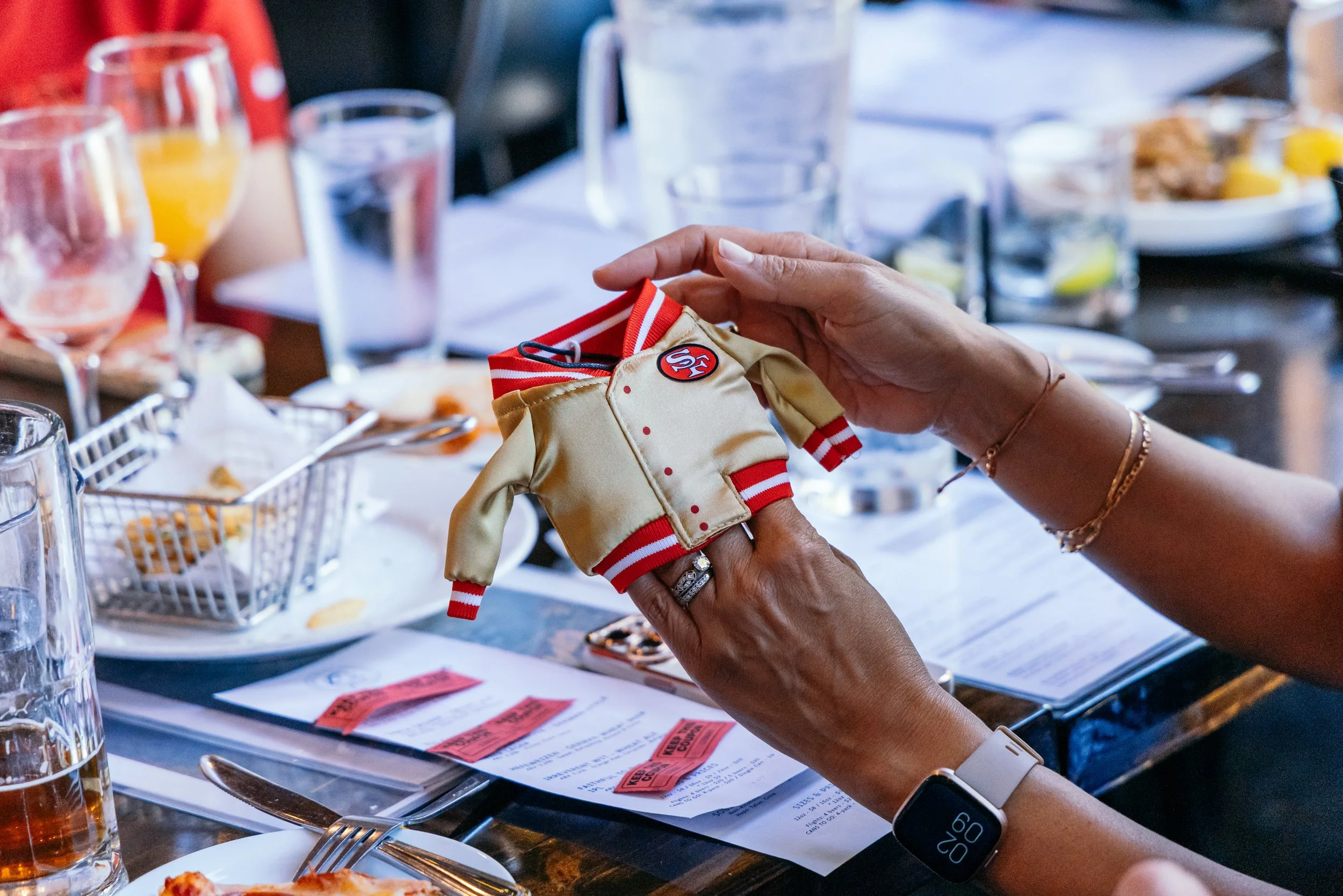 A person holding a small stuffed toy dressed in a beige and red outfit at a restaurant table with glasses of drinks, plates of food, and menus.