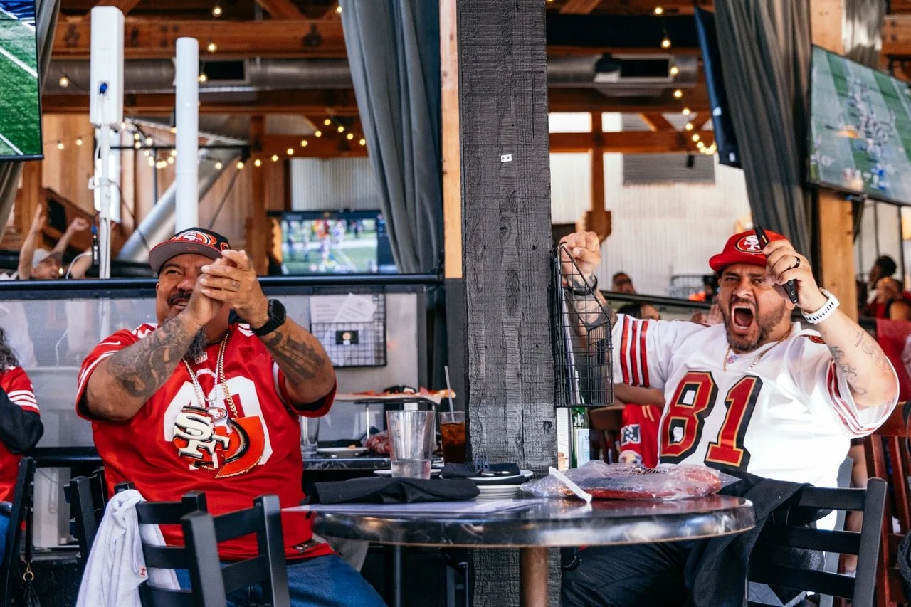 Two men in San Francisco 49ers football jerseys celebrating at a restaurant during a game, with one clapping and the other yelling.