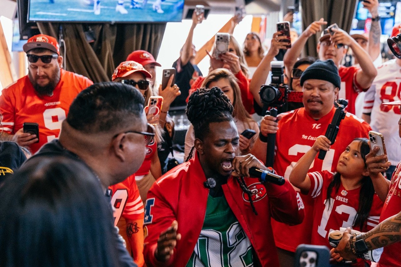 A singer performs passionately at a crowded California 49ers game event, surrounded by fans capturing the moment on their phones.
