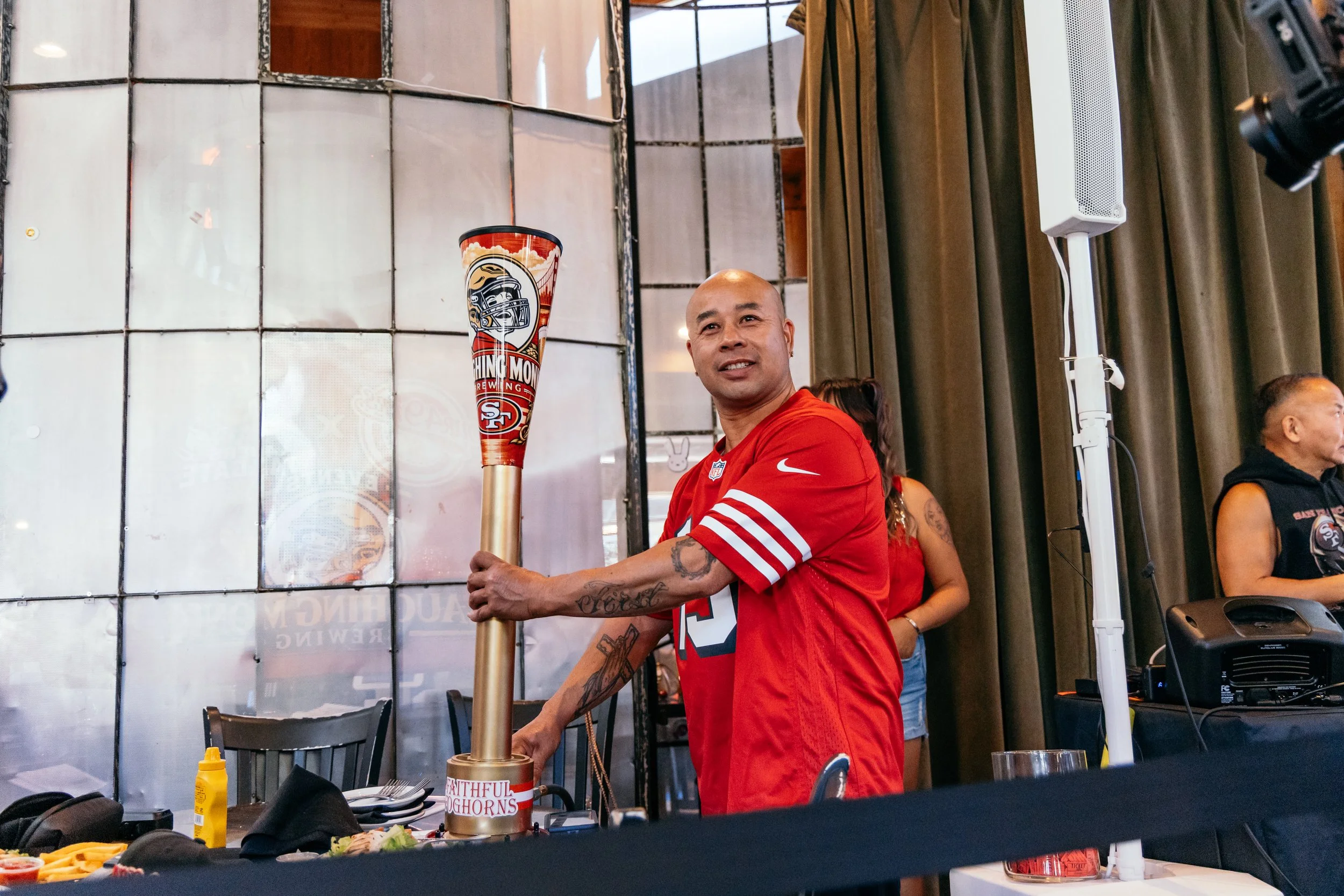 Man in red San Francisco 49ers jersey holding a large Vikings-themed horn at a social event inside a room with curtains and a speaker.
