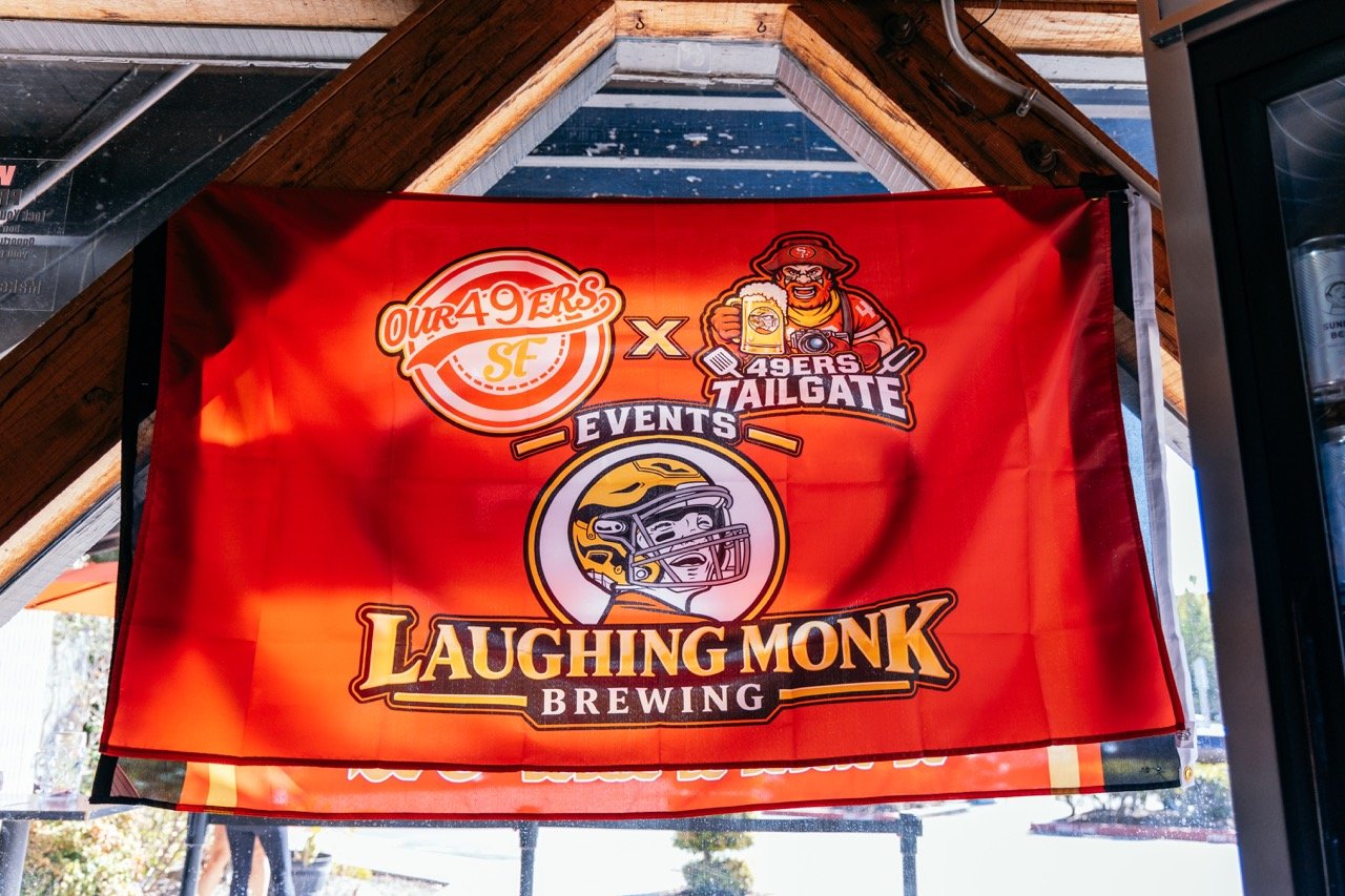A red banner hanging inside a building, displaying the logos of the San Francisco 49ers and Laughing Monk Brewing, promoting a collaborative event.