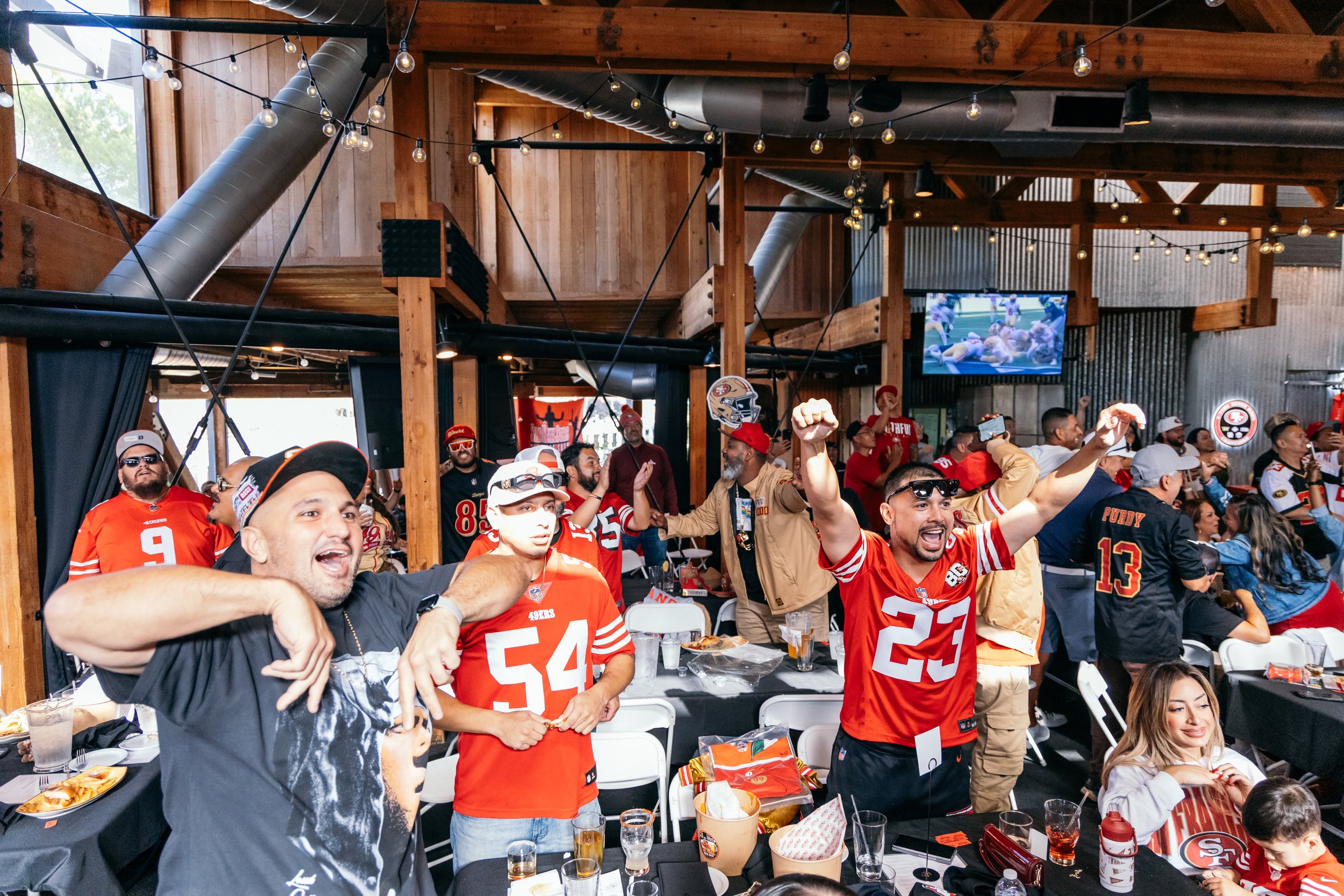 A group of football fans, mostly wearing San Francisco 49ers jerseys and gear, celebrating at an indoor sports bar with wooden interior, string lights, and a large flat-screen TV showing a football game.