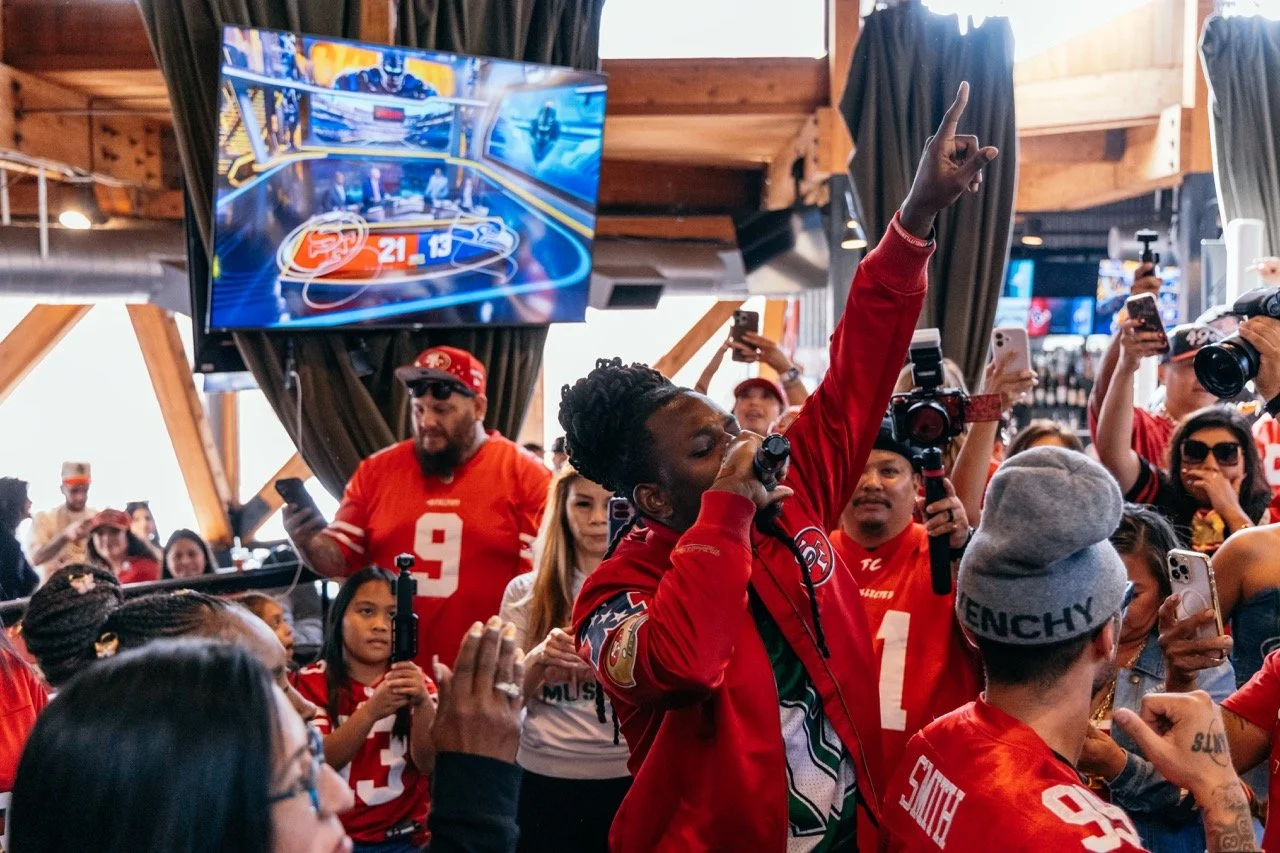 A lively gathering of sports fans, many wearing San Francisco 49ers jerseys and hats, with a woman singing into a microphone in the middle, raising her finger, amid others filming or taking pictures in a wooden, indoor venue with large screens displa
