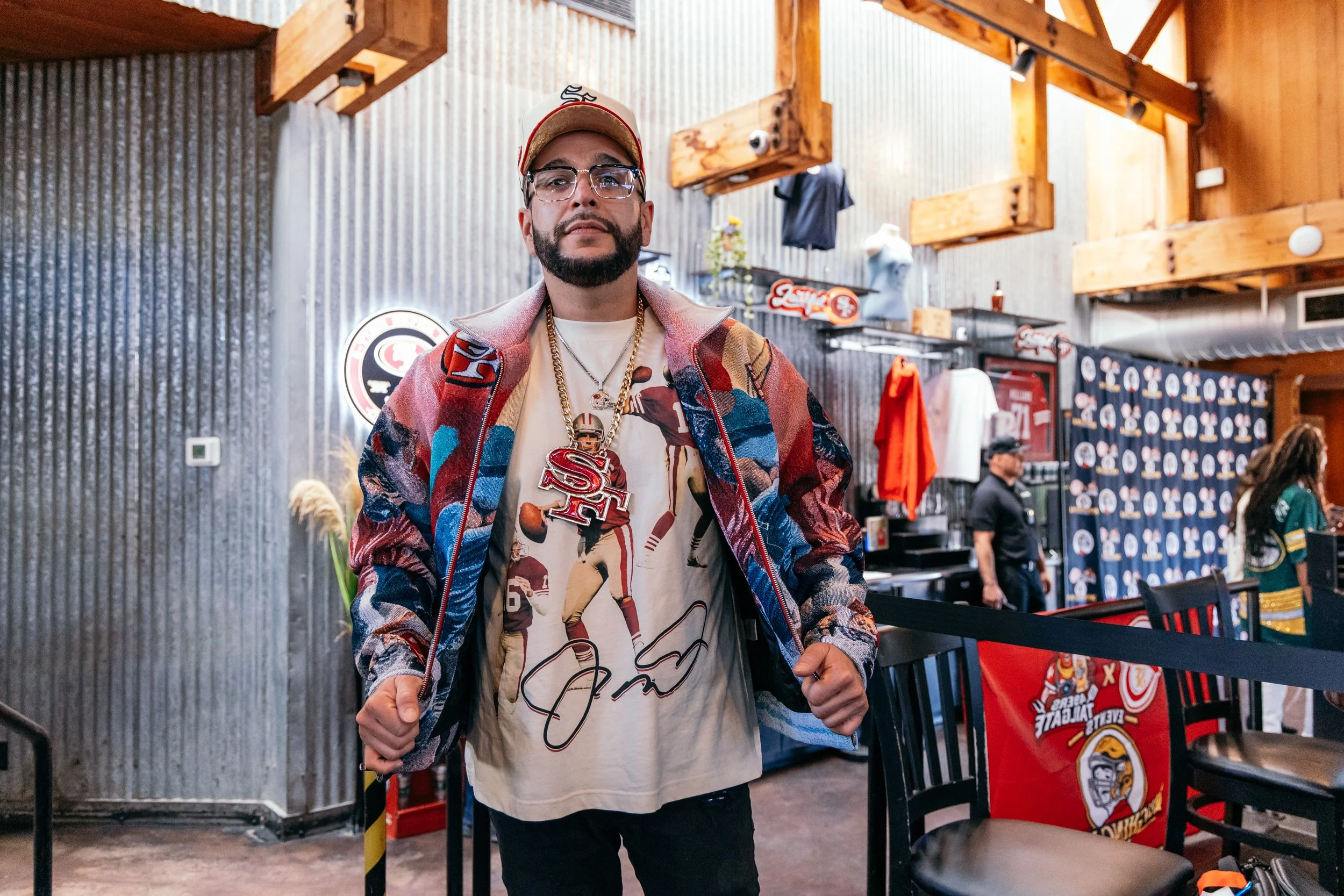 A man wearing glasses, a baseball cap, a colorful jacket, and sports-themed jewelry standing in a restaurant or sports bar decorated with team merchandise and memorabilia.