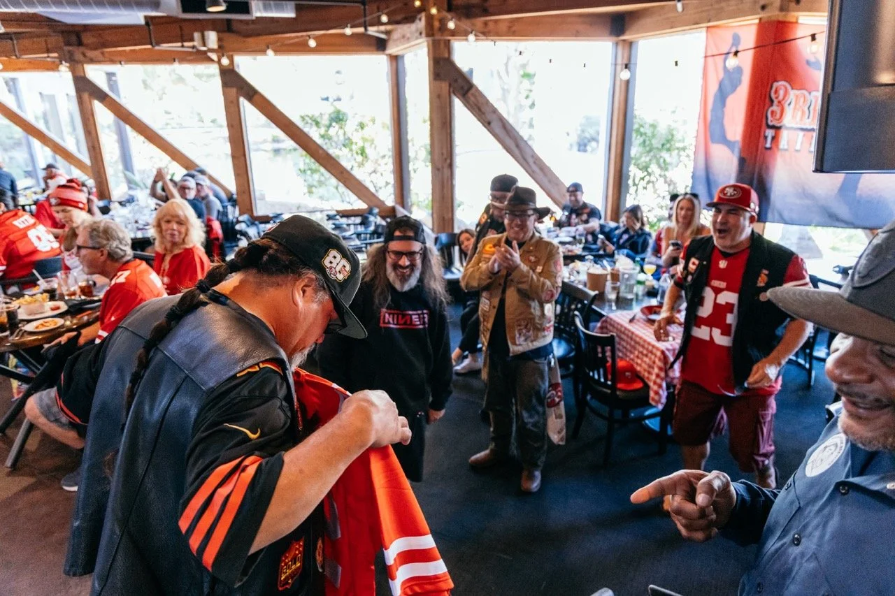 People gathered in a wooden sports bar or restaurant, many wearing San Francisco 49ers jerseys and hats, socializing, and taking photos during a game day celebration.