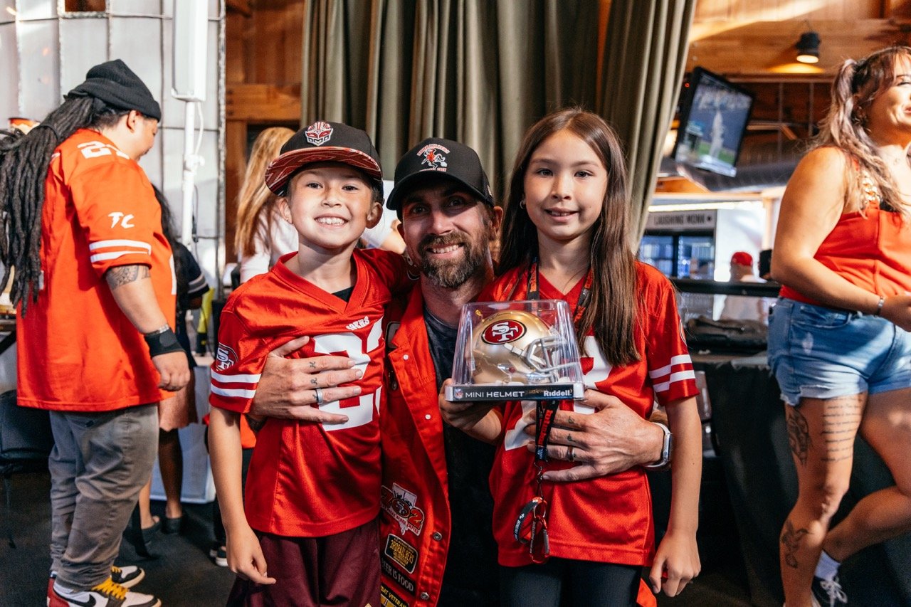 A man and two children dressed in San Francisco 49ers jerseys, smiling and posing for a photo. The girl is holding a mini football helmet. Other people in 49ers attire are in the background at a festive indoor event.
