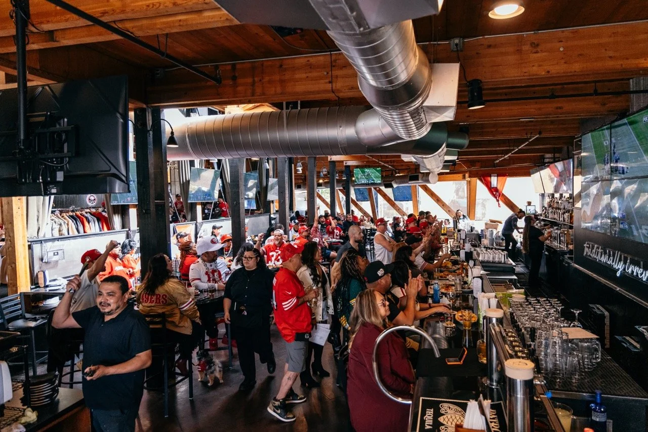 Inside a sports bar with many football fans wearing team jerseys and hats, watching a game on multiple large screens, with some people standing and others sitting at the bar and tables, wooden ceiling and large ventilation ducts visible.
