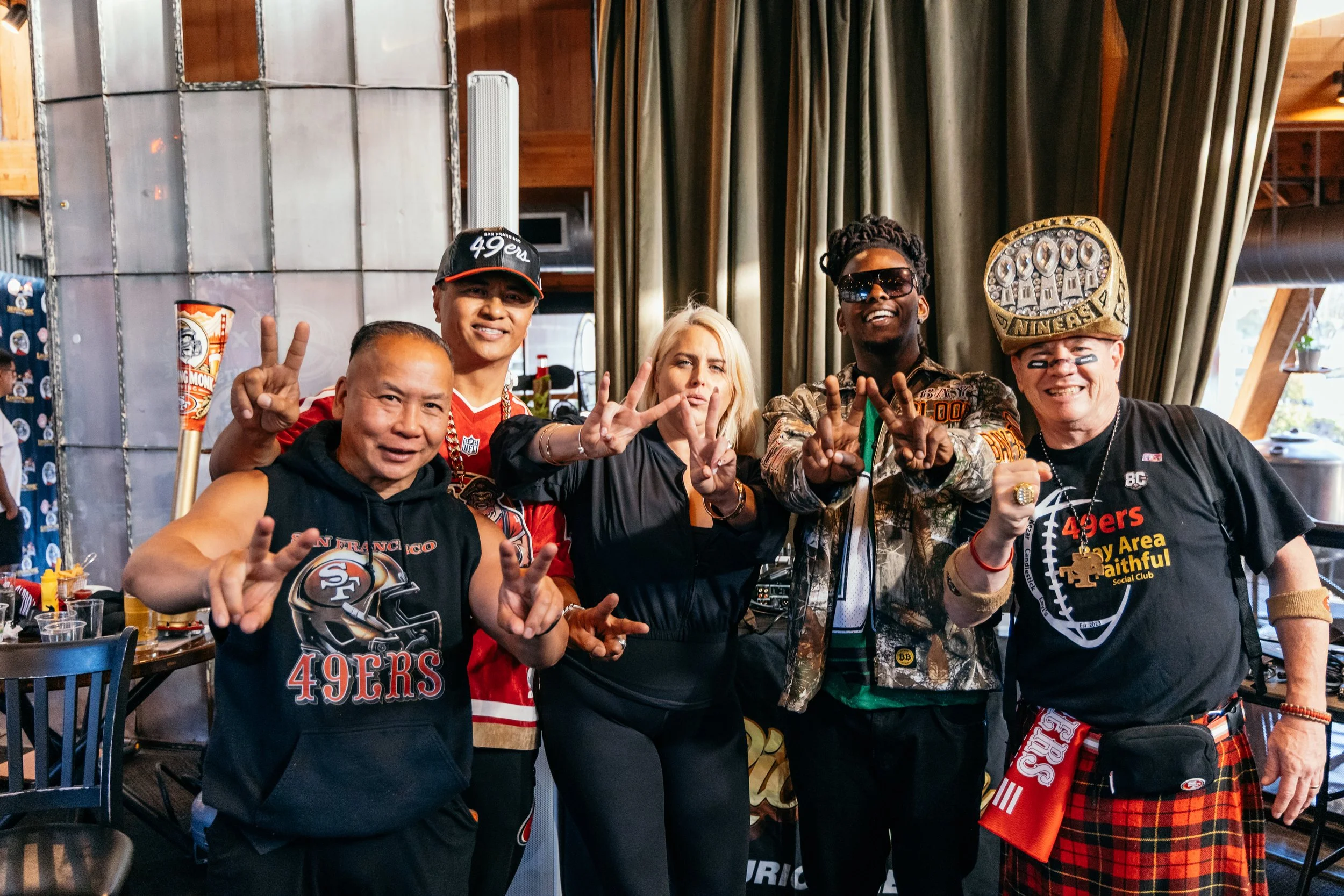A group of five people are posing together indoors, smiling and making hand gestures. They are wearing sports-themed clothing, including a San Francisco 49ers jersey and hat, and are celebrating. The background shows wooden walls, green curtains, and