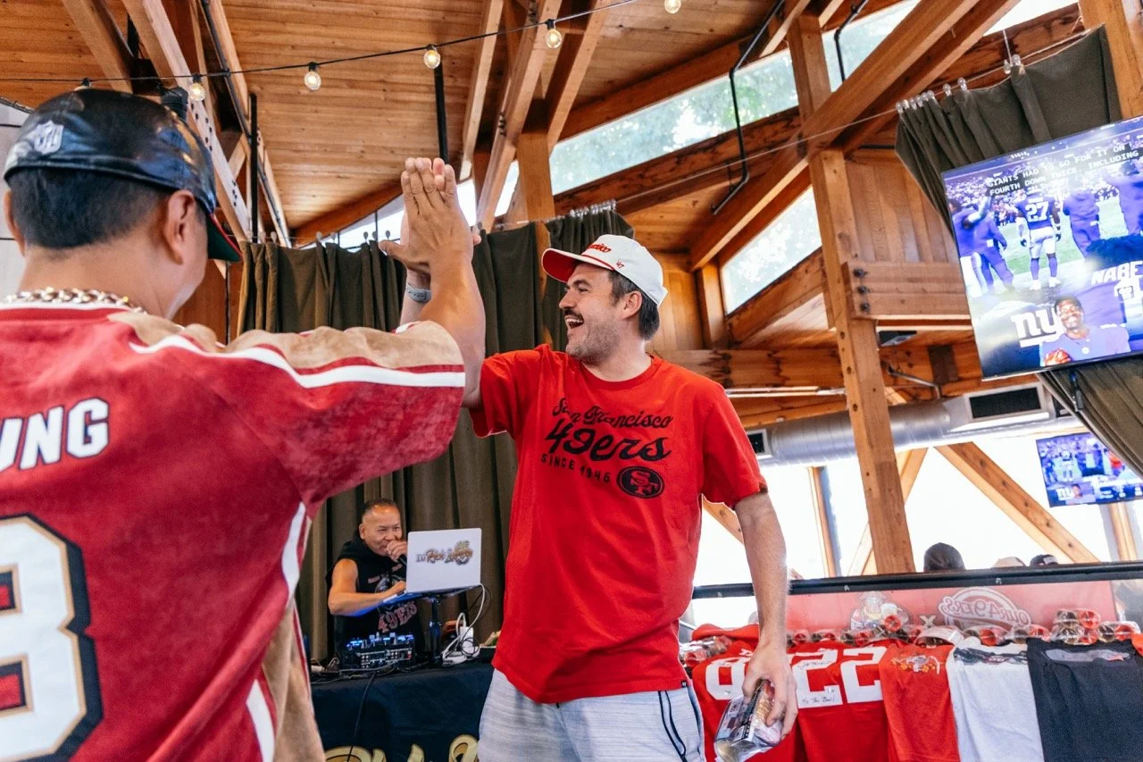 Two men celebrating with a high five at a sports-themed event, with a DJ and a TV screen displaying football games in the background, inside a wooden venue.