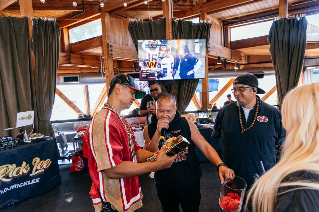 Group of people at an indoor event, with two men and a woman gathered, one man holding a device and another holding a container. In the background, there's a DJ station with equipment and a large TV screen displaying a sports game and graphics.