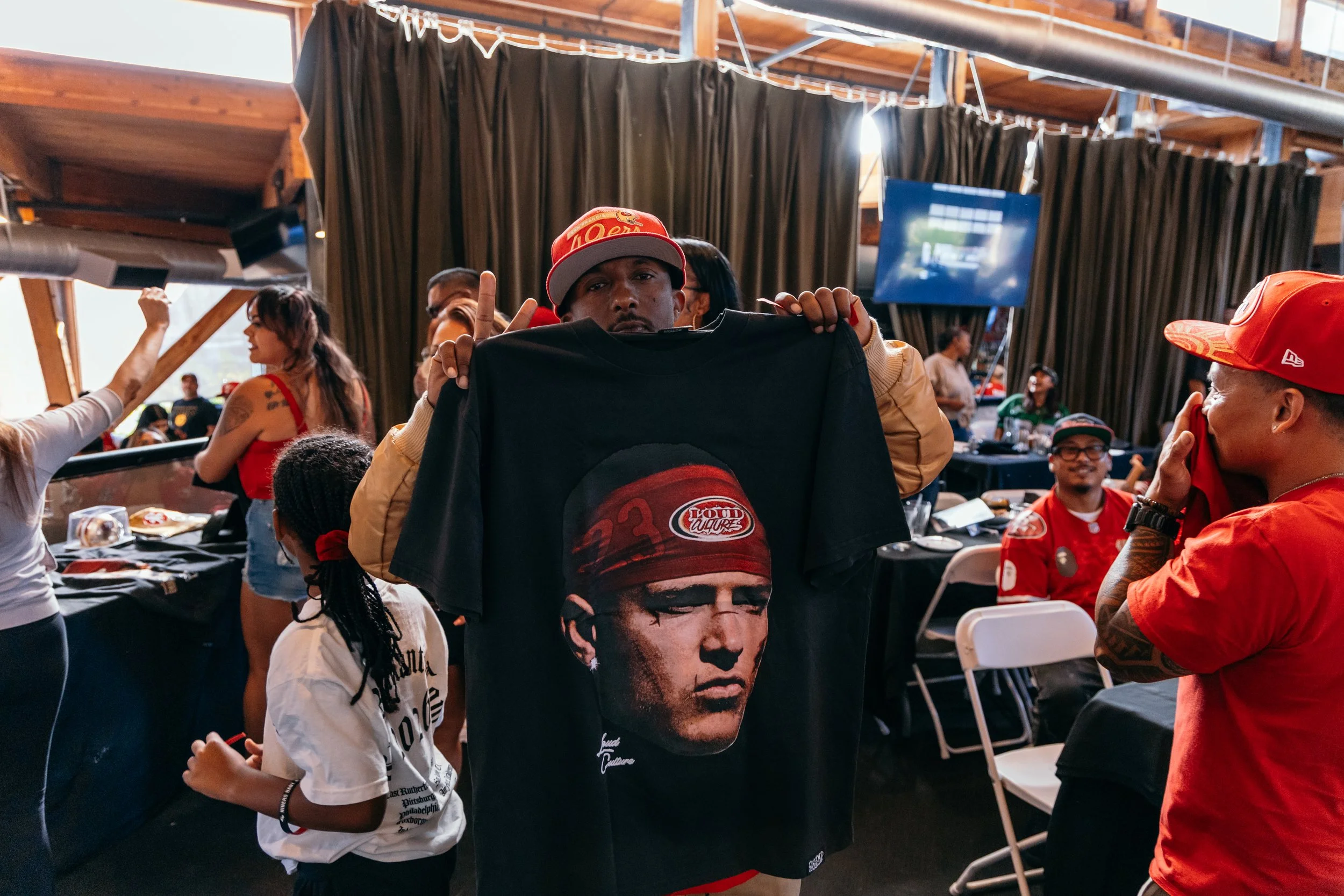Person holding up a t-shirt with a portrait of a man wearing a cap, surrounded by people at an indoor event.