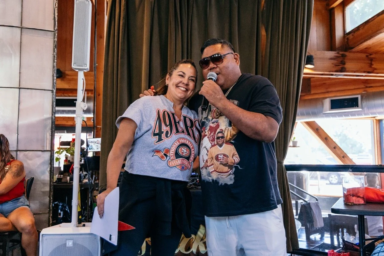 A woman wearing a San Francisco 49ers t-shirt smiling and posing next to a man in sunglasses with a microphone in hand, both standing near a stage or performance area with curtains and large windows behind them.