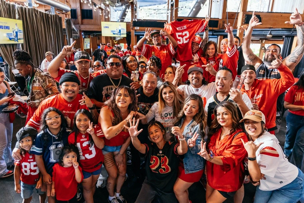 A large group of people wearing San Francisco 49ers football team apparel gathered at a celebration, smiling and posing for the camera inside a decorated wooden venue.