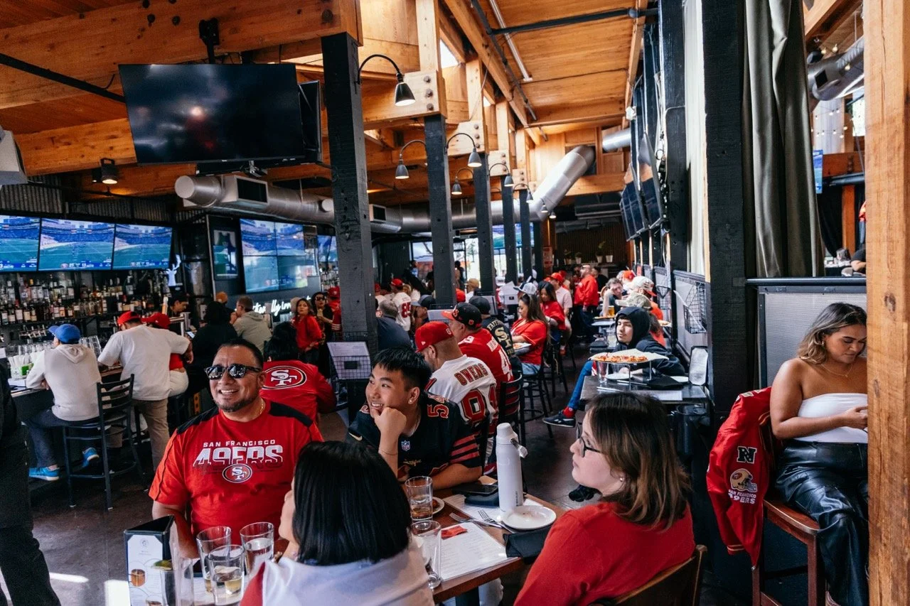 People watching a game at a sports bar decorated with San Francisco 49ers football memorabilia, with multiple TV screens showing a sports event.