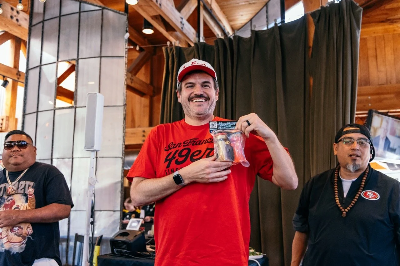A smiling man in a red San Francisco 49ers T-shirt and cap holding a prize in a plastic bag at an indoor event, with two men standing beside him.