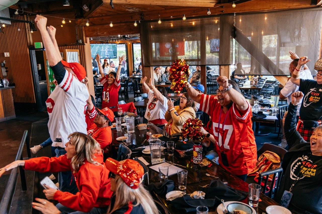 A group of football fans wearing San Francisco 49ers jerseys celebrating and cheering at a restaurant or bar.