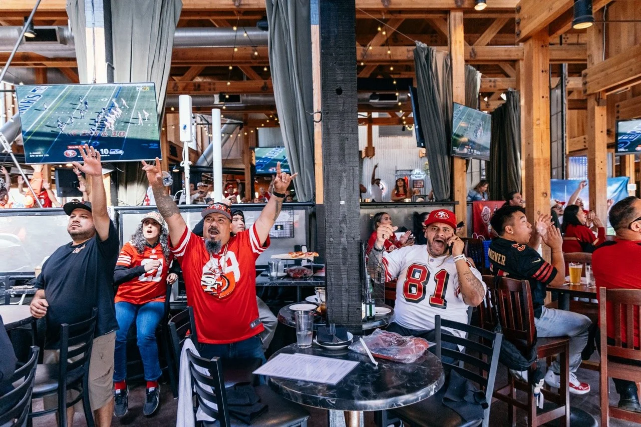 Fans watching a football game on TV at a sports bar, some celebrating, others cheering, in a wooden-finished venue.