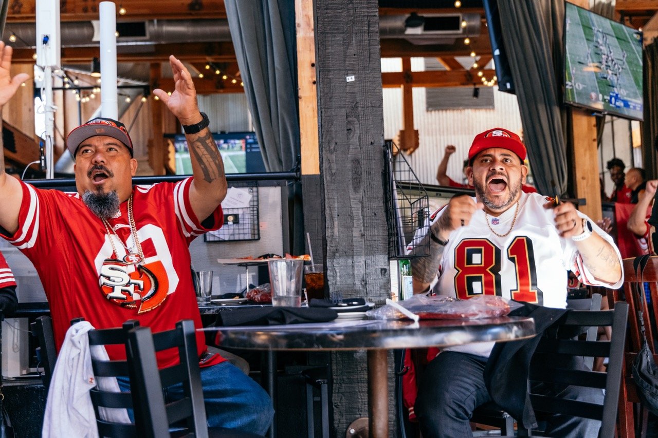Two men wearing San Francisco 49ers football jerseys celebrating enthusiastically at a sports bar or restaurant, with one raising his arms and the other clenching his fists.