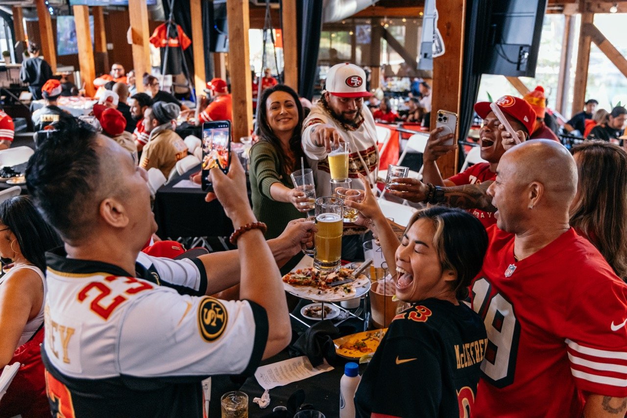 Group of diverse sports fans celebrating and cheering at a restaurant, wearing San Francisco 49ers jerseys and hats, with food and drinks on the table.