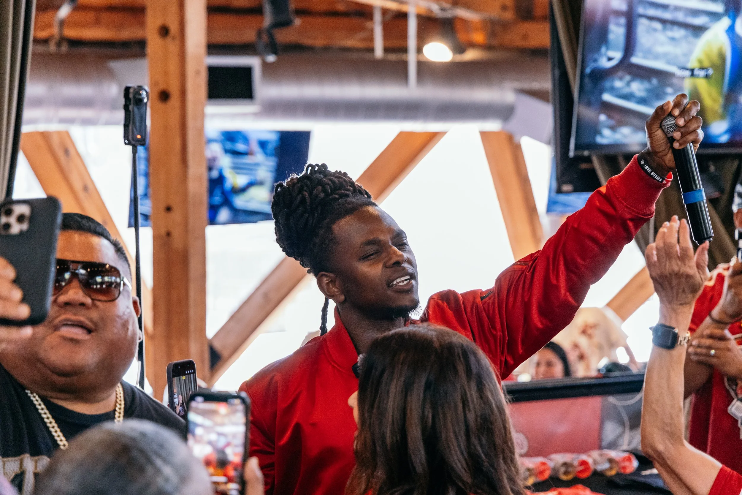 A group of people at a crowded indoor event, some taking photos or videos with their phones. A woman in the center wearing a red jacket is raising her hand, possibly waving or signaling. The setting has wooden beams and a large screen or monitor in t