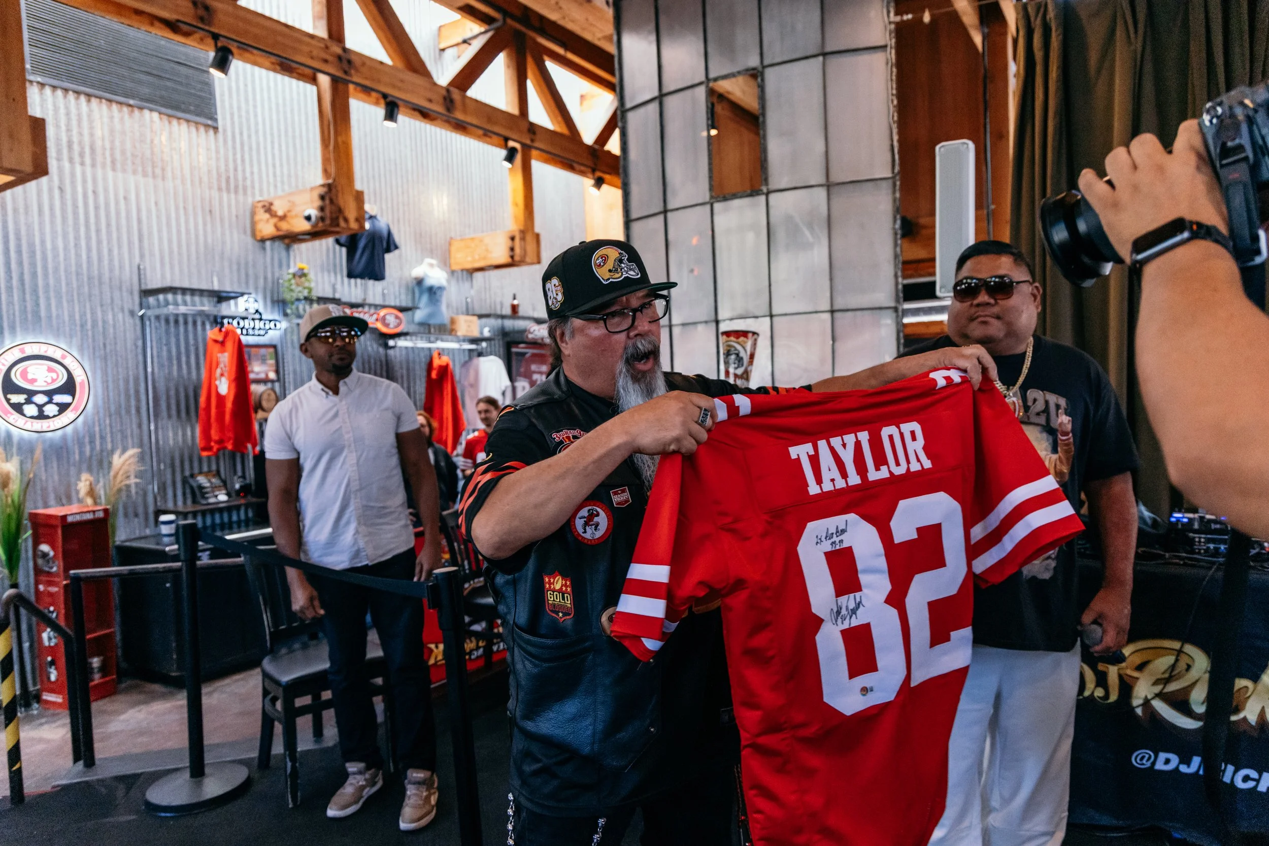 A man wearing glasses and a black cap with a football helmet logo holds and examines a red football jersey with the name 'TAYLOR' and the number 20 during a gathering in a rustic indoor setting with wooden beams and metal siding.