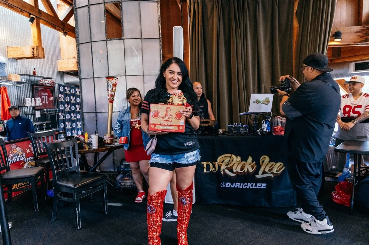 A woman wearing San Francisco 49ers-themed red, black, and silver boots holding a box of Chinese dumplings at a celebration event. She is smiling and standing in front of a DJ booth with a DJ and others in casual attire. There are American football f