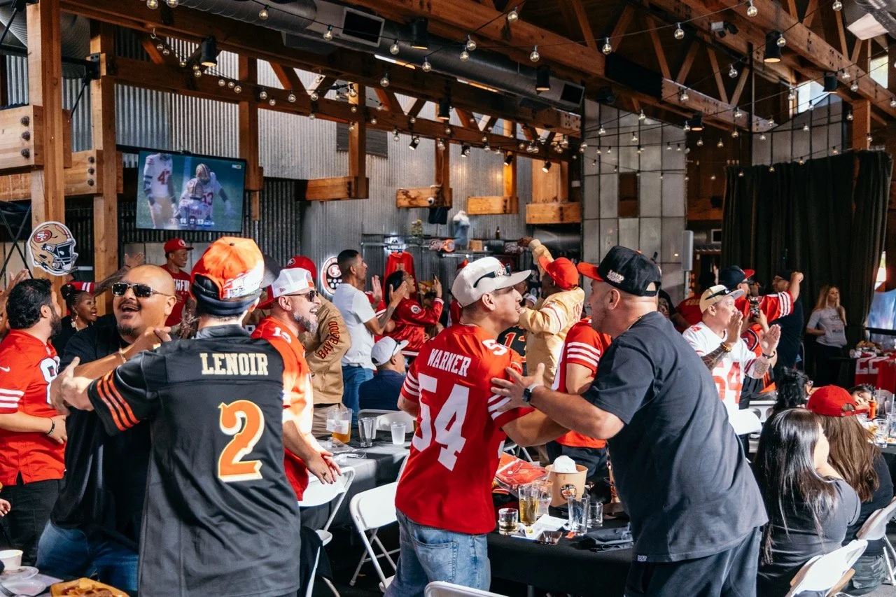 Celebration of San Francisco 49ers fans at a sports bar, cheering and dancing with team jerseys, tattoos, and drinks, while watching a football game on TV.