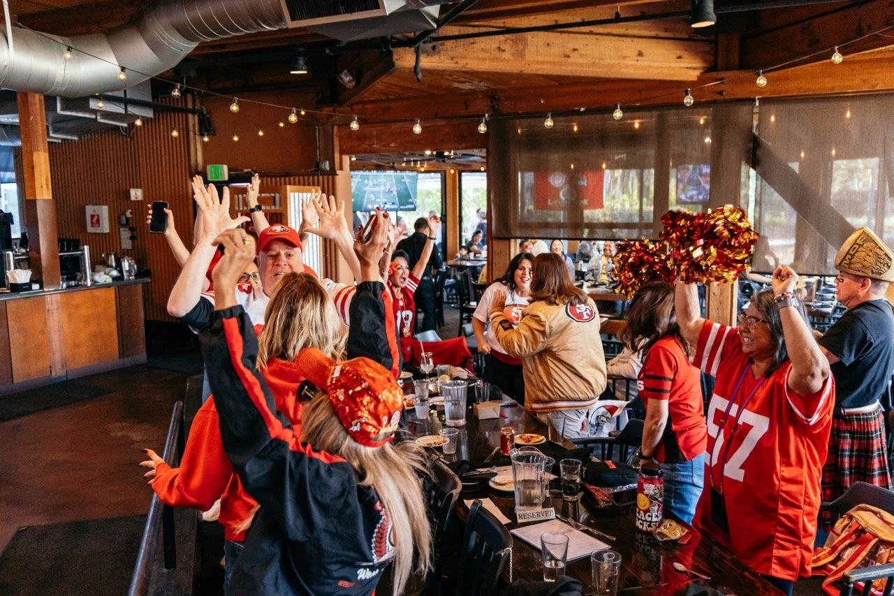 Celebrating San Francisco 49ers fans at a restaurant, wearing team jerseys, hats, and waving pom-poms, with some raising their hands in excitement.