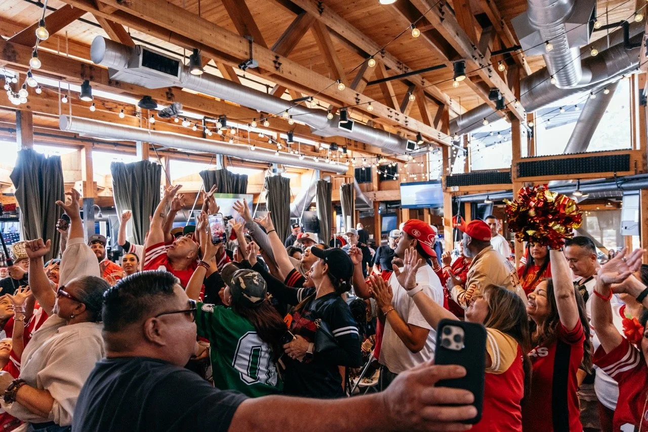 Crowd of people at a lively indoor sports event, some wearing team jerseys and hats, cheering and taking photos inside a wooden venue with string lights and large windows.