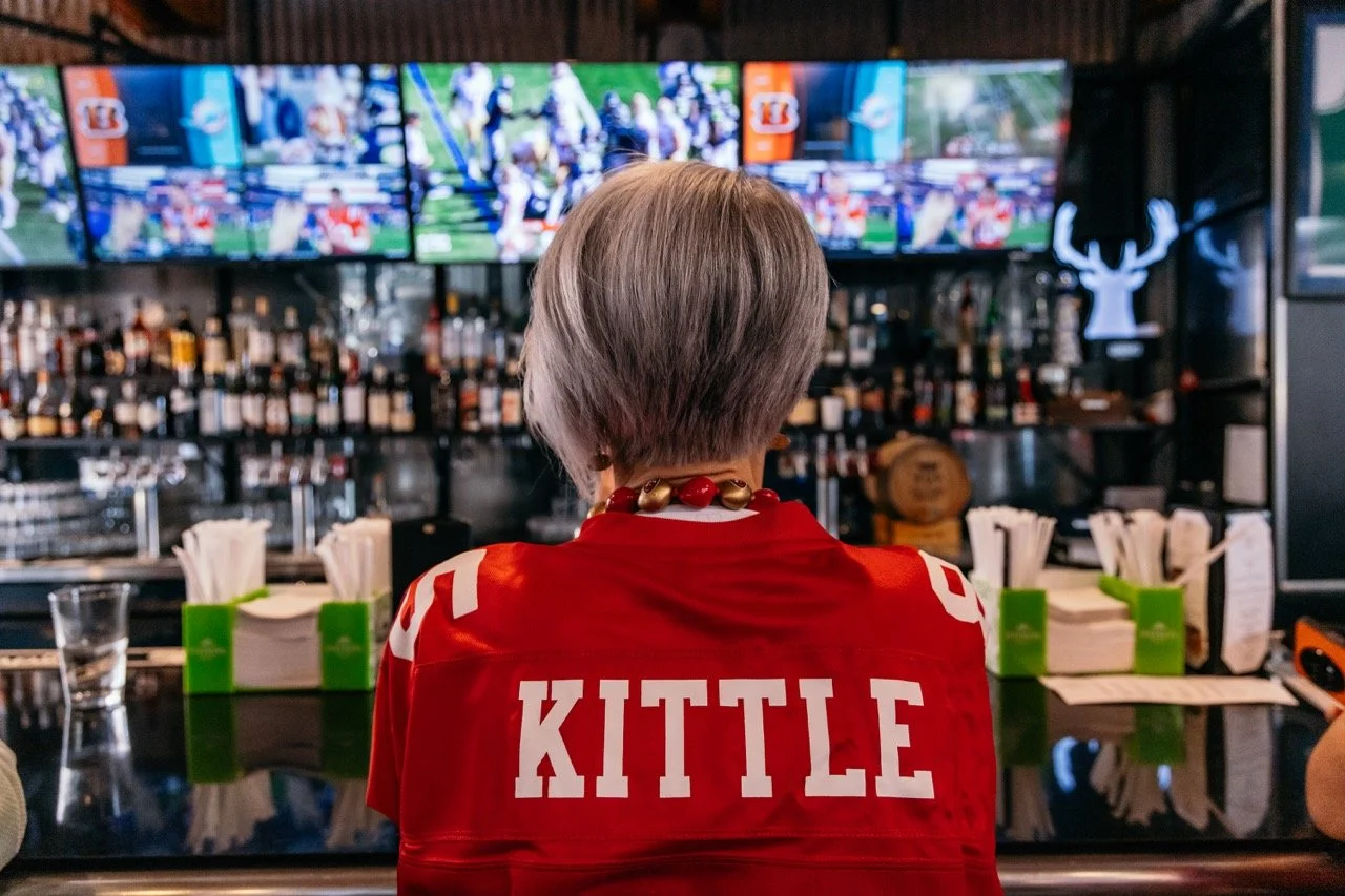 Back view of a person sitting at a bar wearing a red sports jersey with the name KITTLE, watching multiple television screens showing a football game.